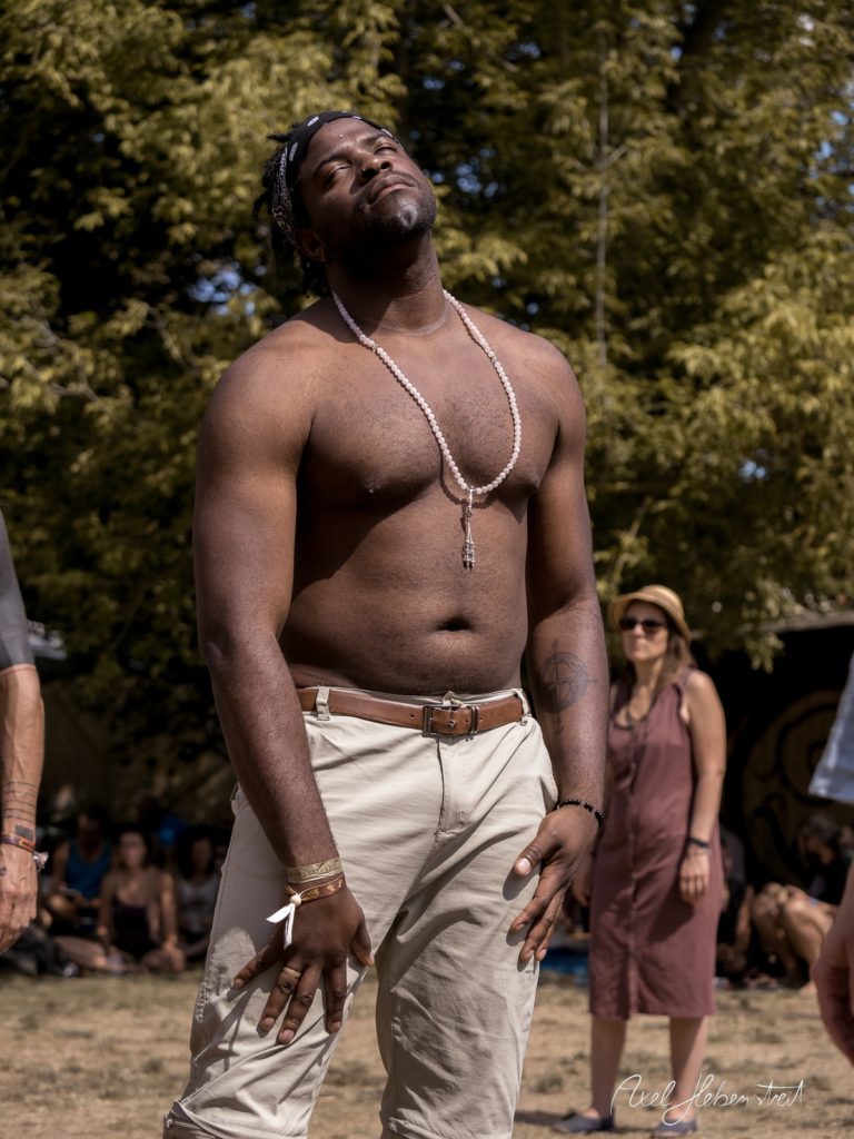 Ein muskulöser Mann mit Perlenkette genießt den Moment in einer Festivalatmosphäre im Freien – © Axel Hebenstreit