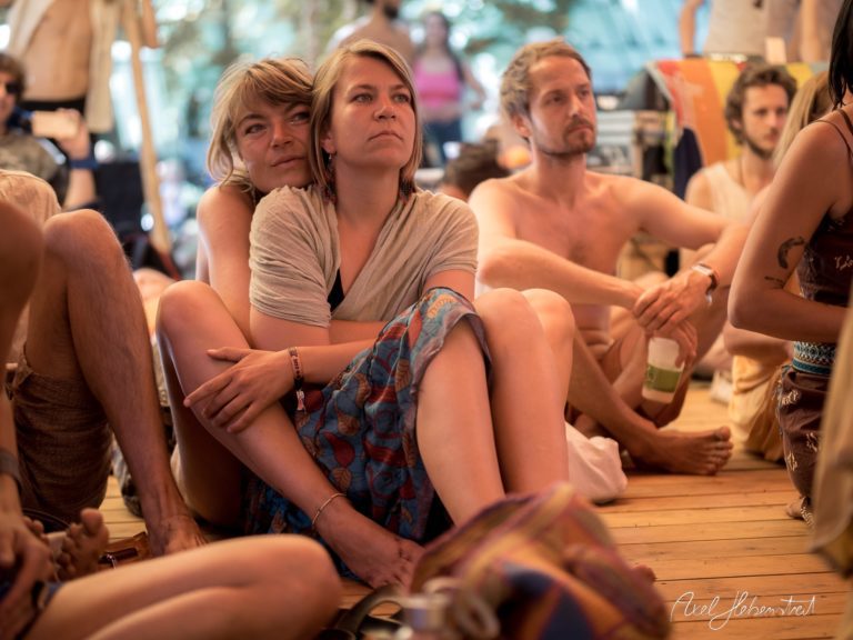 Zwei Frauen sitzen zusammen auf dem Boden eines Festivals und lauschen gebannt einem Vortrag. – © Axel Hebenstreit