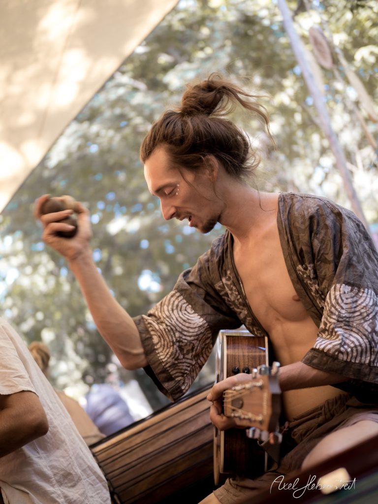 Junger Mann mit Gitarre spielt und schüttelt ein Rasselinstrument, umgeben von natürlicher Festivalkulisse – © Axel Hebenstreit.