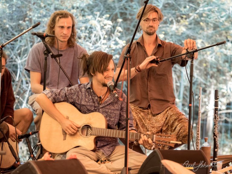 Musiker spielen an einer Bühne, während ein Mann mit Gitarre die entspannte Festivalatmosphäre genießt – © Axel Hebenstreit