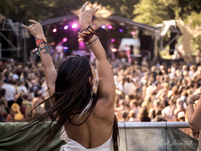 Eine tanzende Person mit langen Haaren hebt die Hände auf einem Festival vor einer lebhaften Bühne – © Axel Hebenstreit