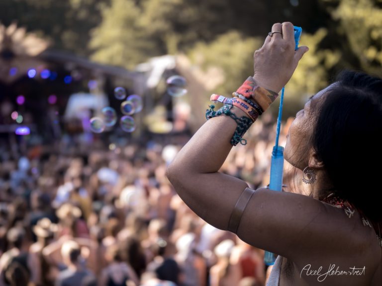 Eine Frau macht ein Foto von einer Festivalbühne, während sie von bunten Seifenblasen umgeben ist – © Axel Hebenstreit