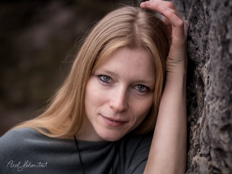 Porträt einer rothaarigen Frau mit sanftem Blick, lehnend an einer rauen Steinwand – © Axel Hebenstreit