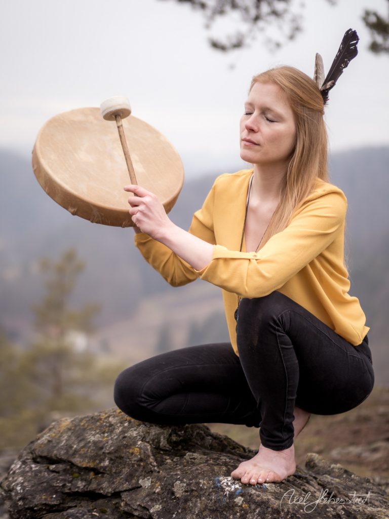 Rothaarige Frau in gelbem Hemd mit Federn im Haar hält eine Trommel auf einem Felsen in bergiger Landschaft – © Axel Hebenstreit