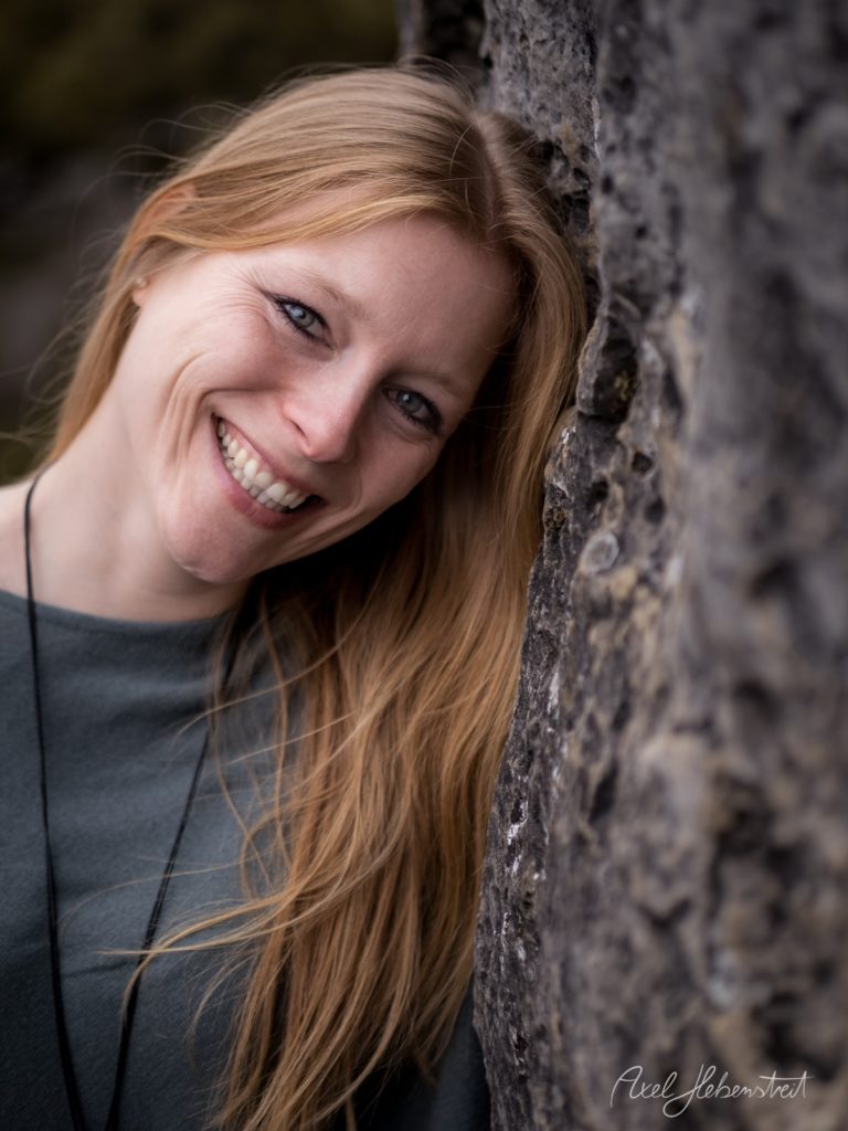 Porträt einer lächelnden Frau mit rötlichem Haar, die hinter einer rauen Steinmauer hervorschaut – © Axel Hebenstreit