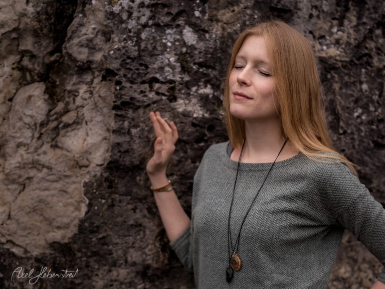 Rothaarige Frau in grauem Pullover lehnt nachdenklich an einer rauen Steinmauer – © Axel Hebenstreit