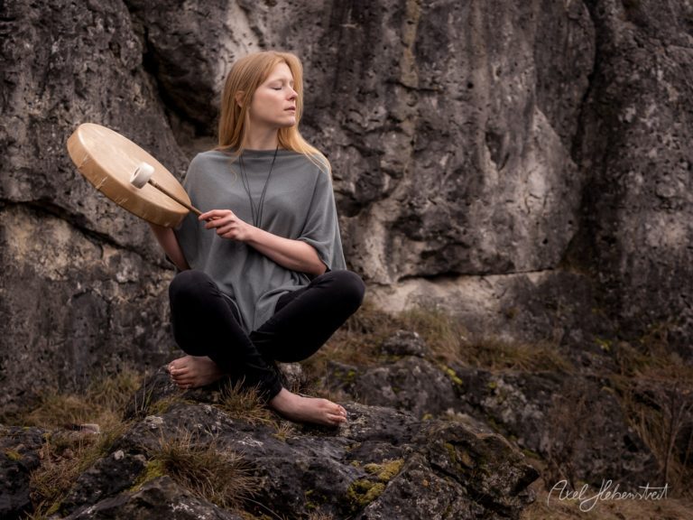 Rothaarige Frau mit Feder sitzt meditativ vor massiven Steinblöcken in naturbelassener Umgebung – © Axel Hebenstreit