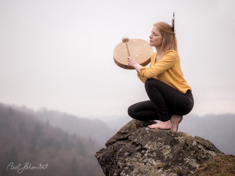 Frau mit rötlichen Haaren in gelbem Oberteil hält Schamanentrommel auf Berggipfel mit nebliger Landschaft – © Axel Hebenstreit