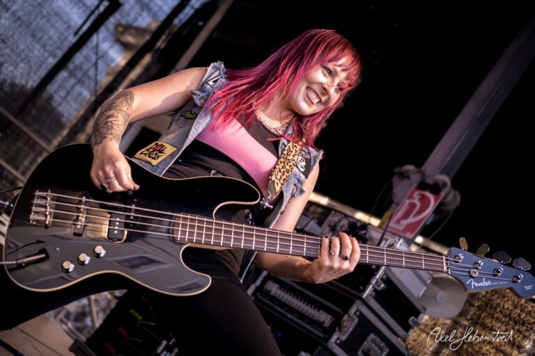 Dynamische Bassistin mit pinken Haaren spielt energisch auf einer Bühne während eines Konzerts – © Axel Hebenstreit