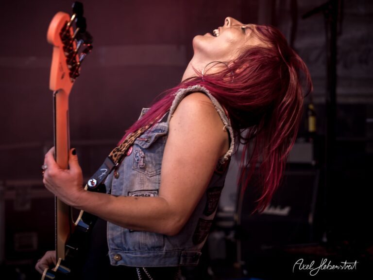 Eine Musikerin mit pinken Haaren spielt begeistert E-Gitarre auf einer Bühne während eines Konzerts – © Axel Hebenstreit