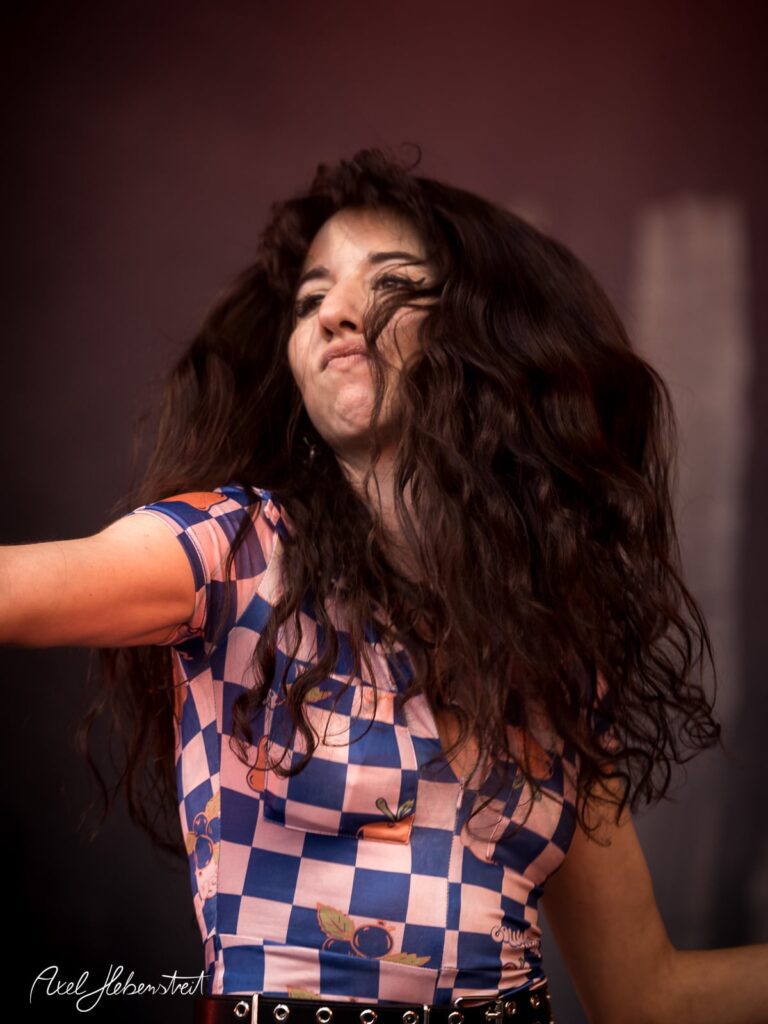 Eine energiegeladene Sängerin mit lockigem Haar, die bei einem Konzert leidenschaftlich performt – © Axel Hebenstreit