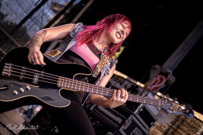 Energiegeladene Musikerin mit pinken Haaren, die leidenschaftlich Bass auf der Bühne spielt – © Axel Hebenstreit