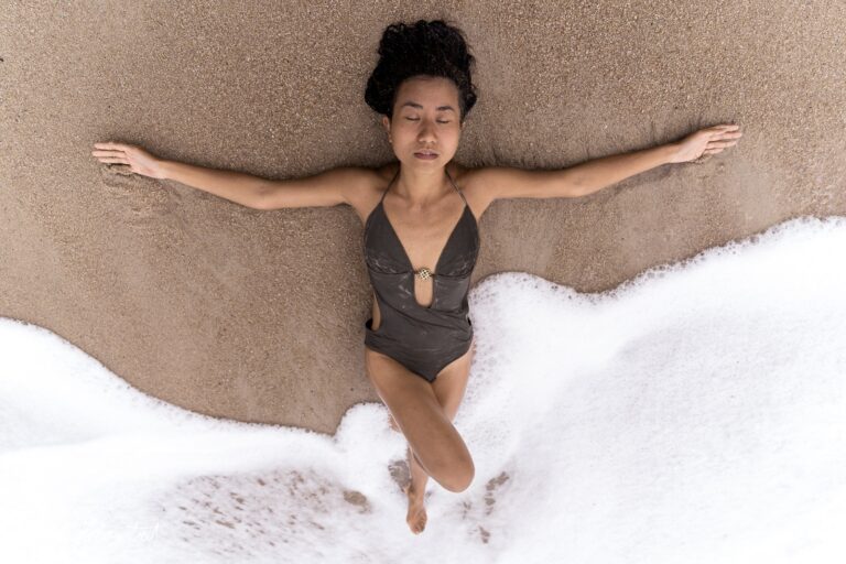 Eine Person in einem Braunton-Badeanzug liegt entspannt im Sand, während Wellen sanft um sie herumspülen – © Axel Hebenstreit.