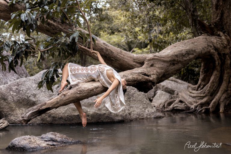 Eine Person in einem fließenden Outfit entspannt auf einem Baumstamm über einem Gewässer in einer natürlichen Umgebung – © Axel Hebenstreit.