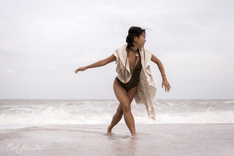 Eine Frau in einem Badeanzug und einem Poncho tanzt fröhlich am Strand mit dramatischen Wellen im Hintergrund – © Axel Hebenstreit