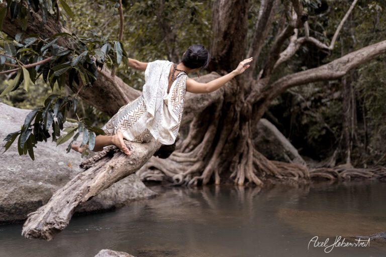 Frau im weißen Kleid balanciert auf einem Baumstamm über einem ruhigen Gewässer in einer geheimnisvollen Naturkulisse – © Axel Hebenstreit