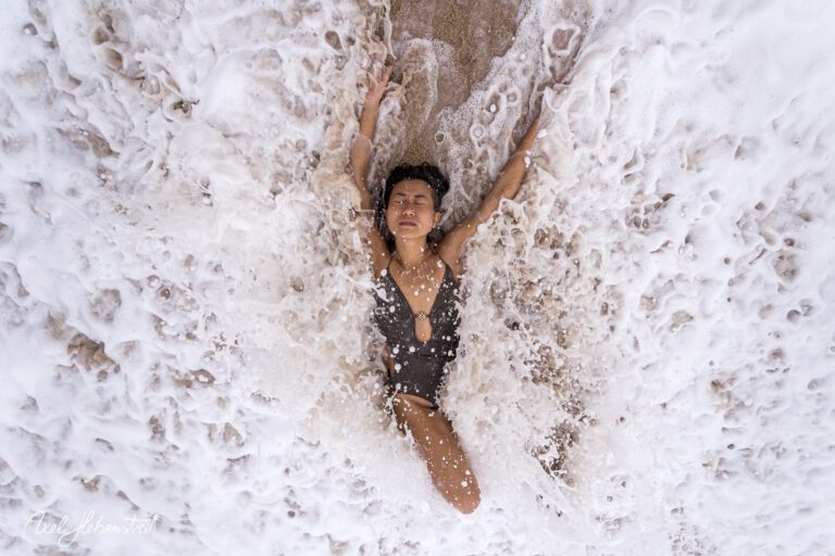 Eine Frau liegt im Schaum des Wassers am Strand, während die Wellen sanft um sie herumspielen. – © Axel Hebenstreit