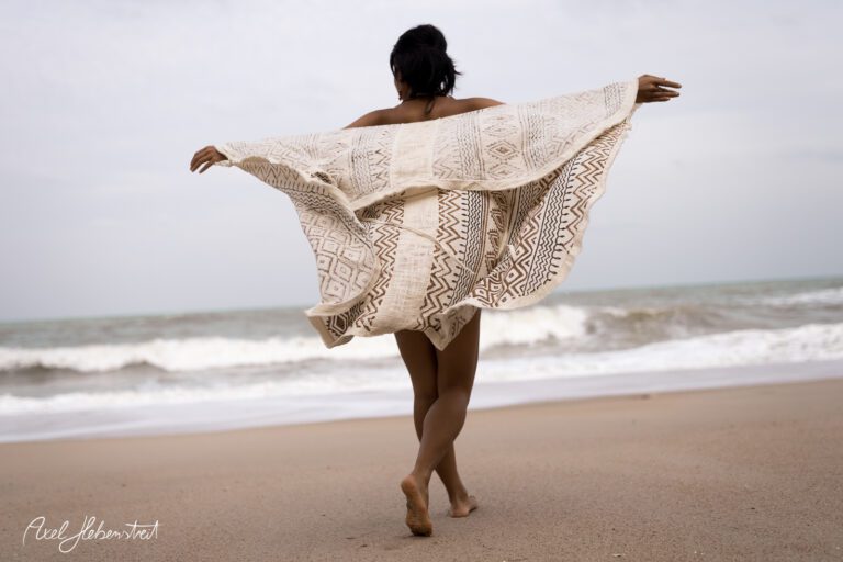 Frau mit einem Tuch in der Hand, die am Strand beim Wasser entlangläuft und leicht dreht – © Axel Hebenstreit