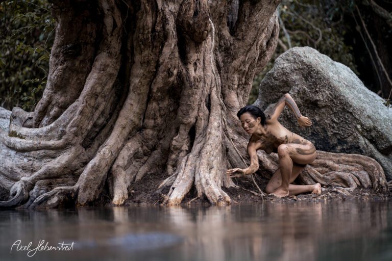 Eine Frau mit Körperbemalung interagiert spielerisch mit dem Wasser eines ruhigen Naturteichs – © Axel Hebenstreit