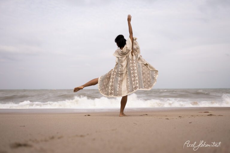 Eine tanzende Frau in einem gemusterten Kleid steht barfuß am Strand und genießt die Meeresbrise – © Axel Hebenstreit