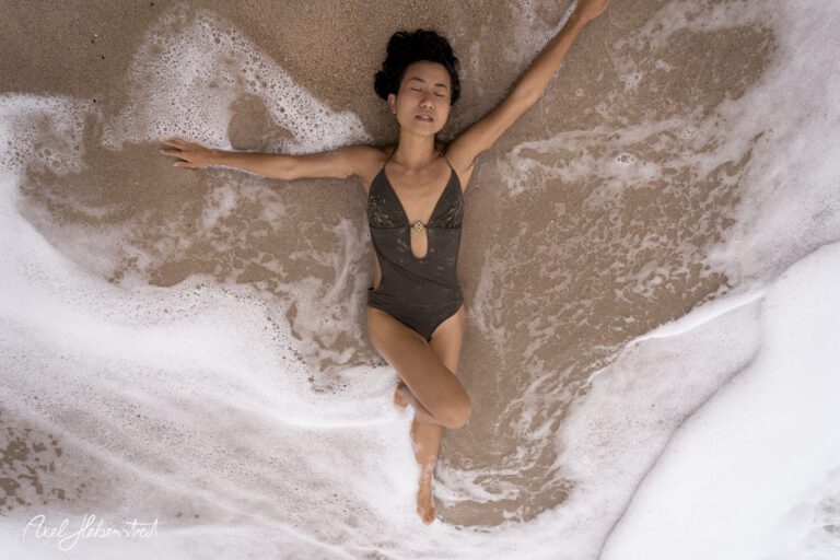 Eine Frau in einem braunen Badeanzug liegt entspannt im sandigen Wasser und genießt die Wellen. – © Axel Hebenstreit