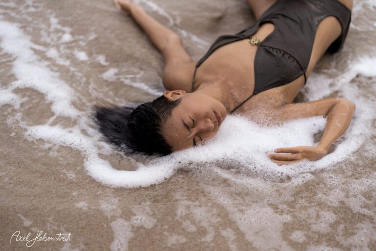 Eine Person liegt entspannt am Strand im Wasser, umgeben von Schaum und einer ruhigen Atmosphäre – © Axel Hebenstreit.