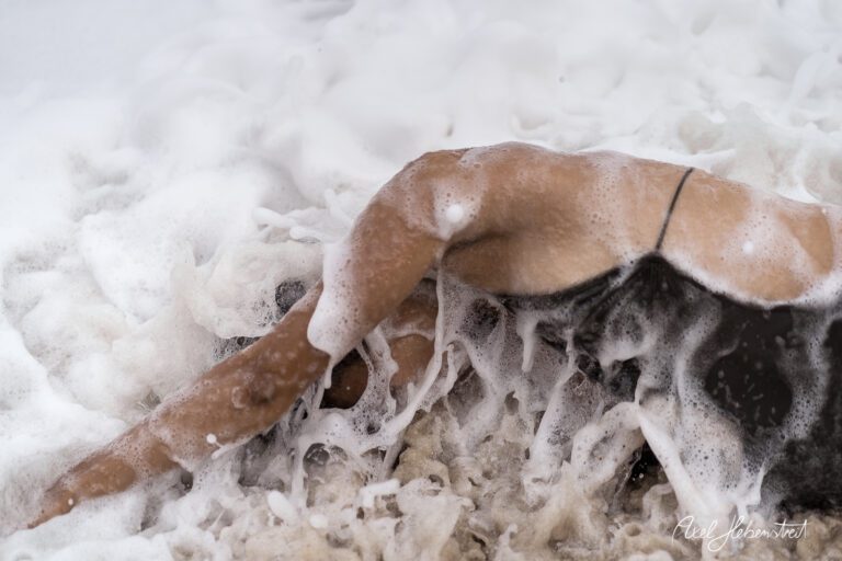 Eine Person liegt im schäumenden Wasser und taucht teilweise in den Schaum ein, umgeben von Sand und Wellen – © Axel Hebenstreit