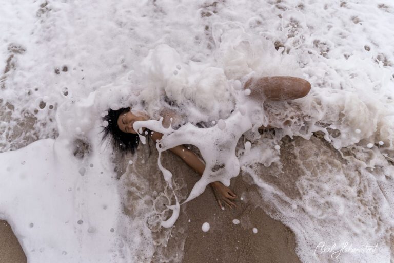 Eine Person liegt am Strand, umgeben von schäumenden Wellen, und vermittelt eine ruhige, träumerische Stimmung. – © Axel Hebenstreit