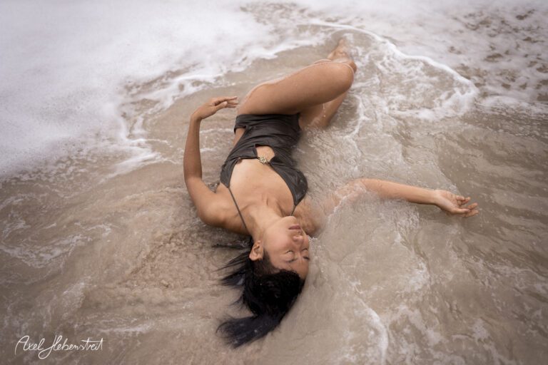 Eine junge Frau liegt entspannt im flachen Wasser am Strand, umgeben von sanften Wellen – © Axel Hebenstreit.