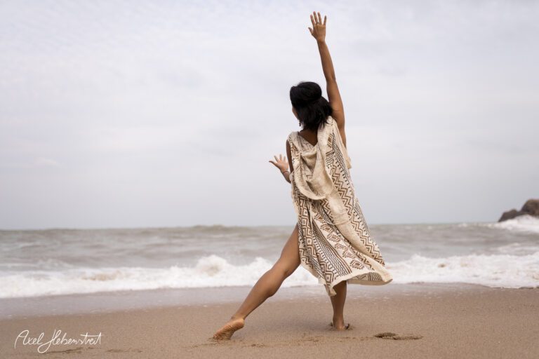 Frau in einem leichten Kleid tanzt spielerisch am Strand mit erhobener Hand – © Axel Hebenstreit