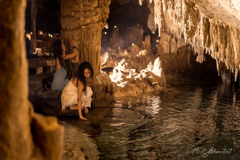 Zwei Frauen in einem Höhlenraum mit Stalaktiten, während eine Wasseroberfläche sanft berührt wird – © Axel Hebenstreit