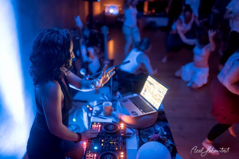 DJ spielt an einem Mischpult, während Tänzer im Hintergrund eine lebhafte Atmosphäre schaffen – © Axel Hebenstreit