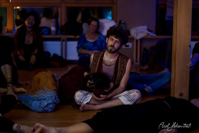 Eine meditative Szene mit Teilnehmern in einem Yogastudio, einer Person umarmt einen anderen auf dem Boden. – © Axel Hebenstreit