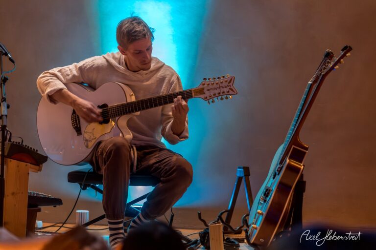 Gitarrist spielt konzentriert auf seiner akustischen Gitarre, umgeben von sanftem Licht in entspannter Atmosphäre – © Axel Hebenstreit