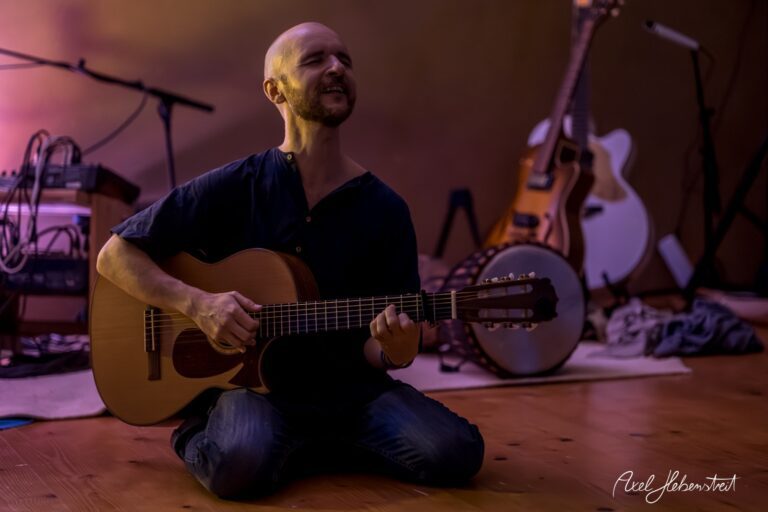 Ein Mann sitzt auf dem Boden und spielt mit Leidenschaft Gitarre in einer stimmungsvollen Umgebung – © Axel Hebenstreit