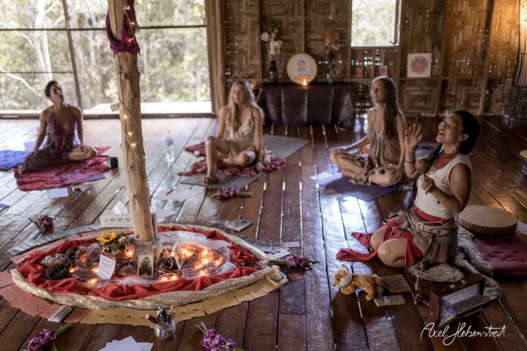 Frauen in meditativer Pose um einen schamanischen Altar in einem Holzraum, umgeben von Natur – © Axel Hebenstreit