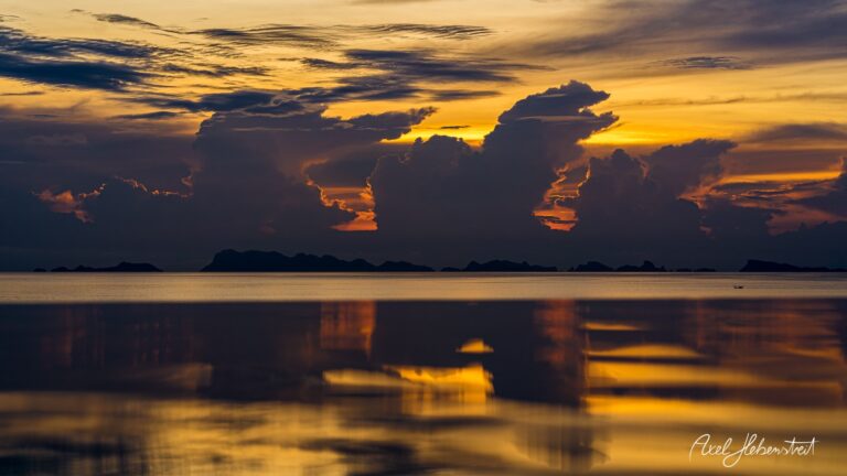 Lebhafte Wolkenformationen spiegeln sich im ruhigen Wasser bei Sonnenuntergang über einer Küstenlandschaft – © Axel Hebenstreit