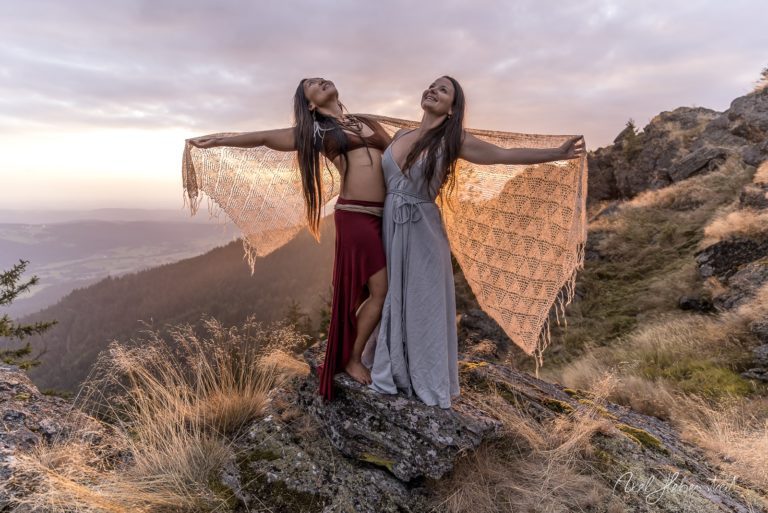 Zwei Frauen stehen auf einem Felsen in der Natur und breiten ihre Arme aus, während die Sonne untergeht. – © Axel Hebenstreit