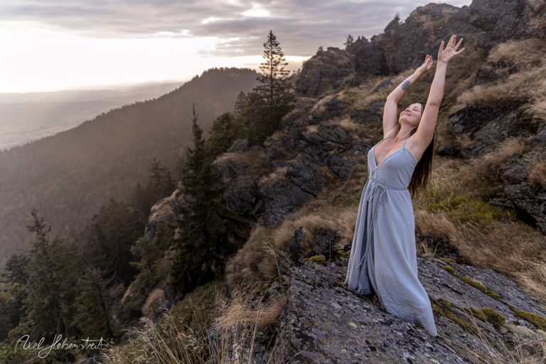 Junge Frau in einem grauen Kleid, die mit ausgebreiteten Armen auf einem Felsen in der Natur steht – © Axel Hebenstreit