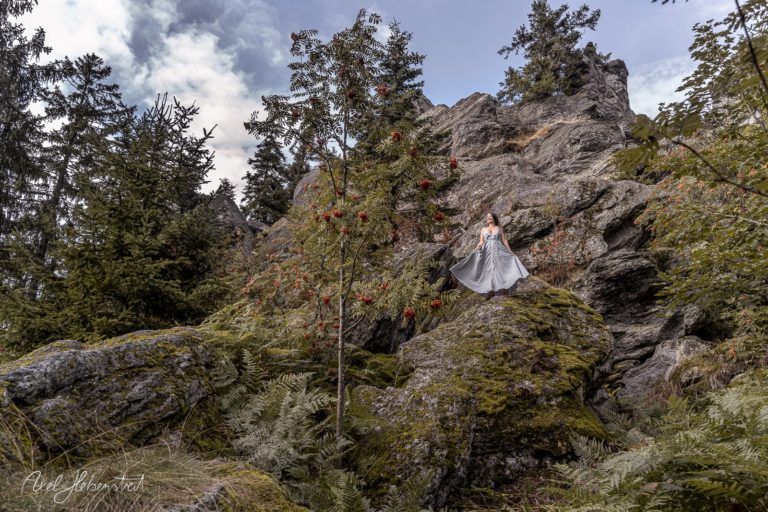 Eine Frau in einem weißen Kleid steht auf einem Felsen in einer bewaldeten Berglandschaft – © Axel Hebenstreit