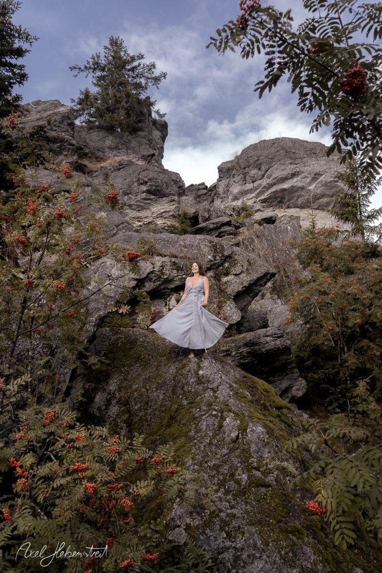 Eine Frau in einem grauen Kleid steht auf einem Felsen umgeben von grünen Pflanzen und roten Beeren – © Axel Hebenstreit