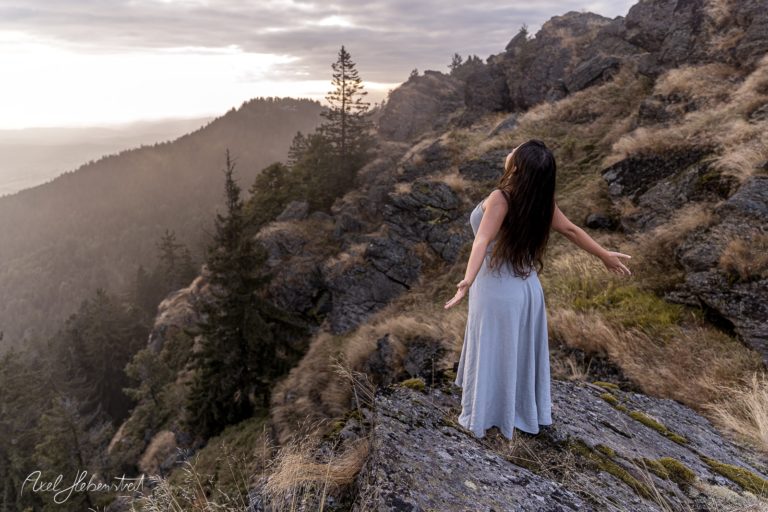 Frau in einem langen Kleid steht auf einem Felsen, breitet die Arme aus und genießt die Aussicht in der Natur – © Axel Hebenstreit