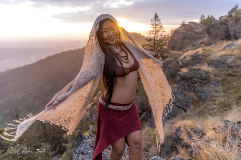 Eine Frau in traditioneller Kleidung tanzt am Rand eines Berges bei Sonnenuntergang. – © Axel Hebenstreit