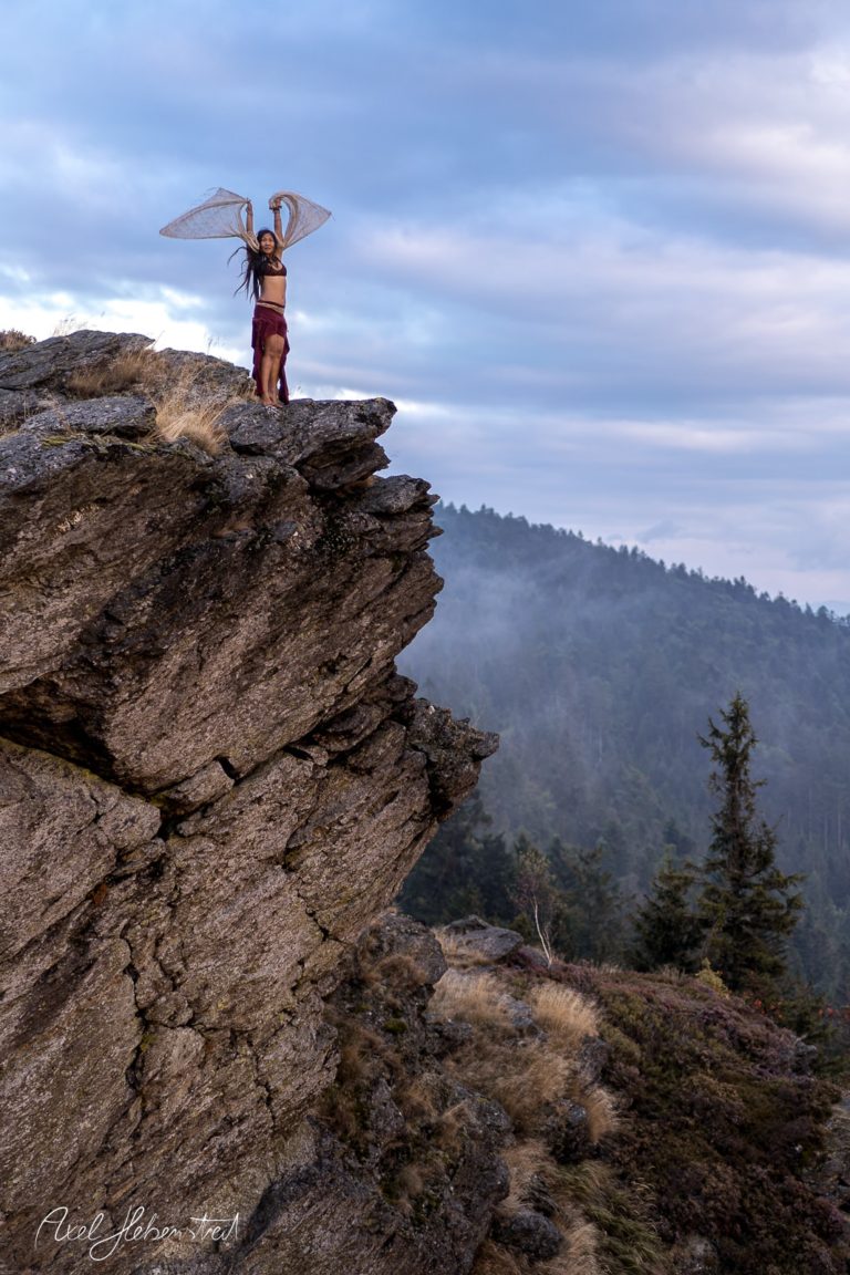 Eine Person mit großen, filigranen Flügeln steht majestätisch auf einem Felsen in der Natur – © Axel Hebenstreit
