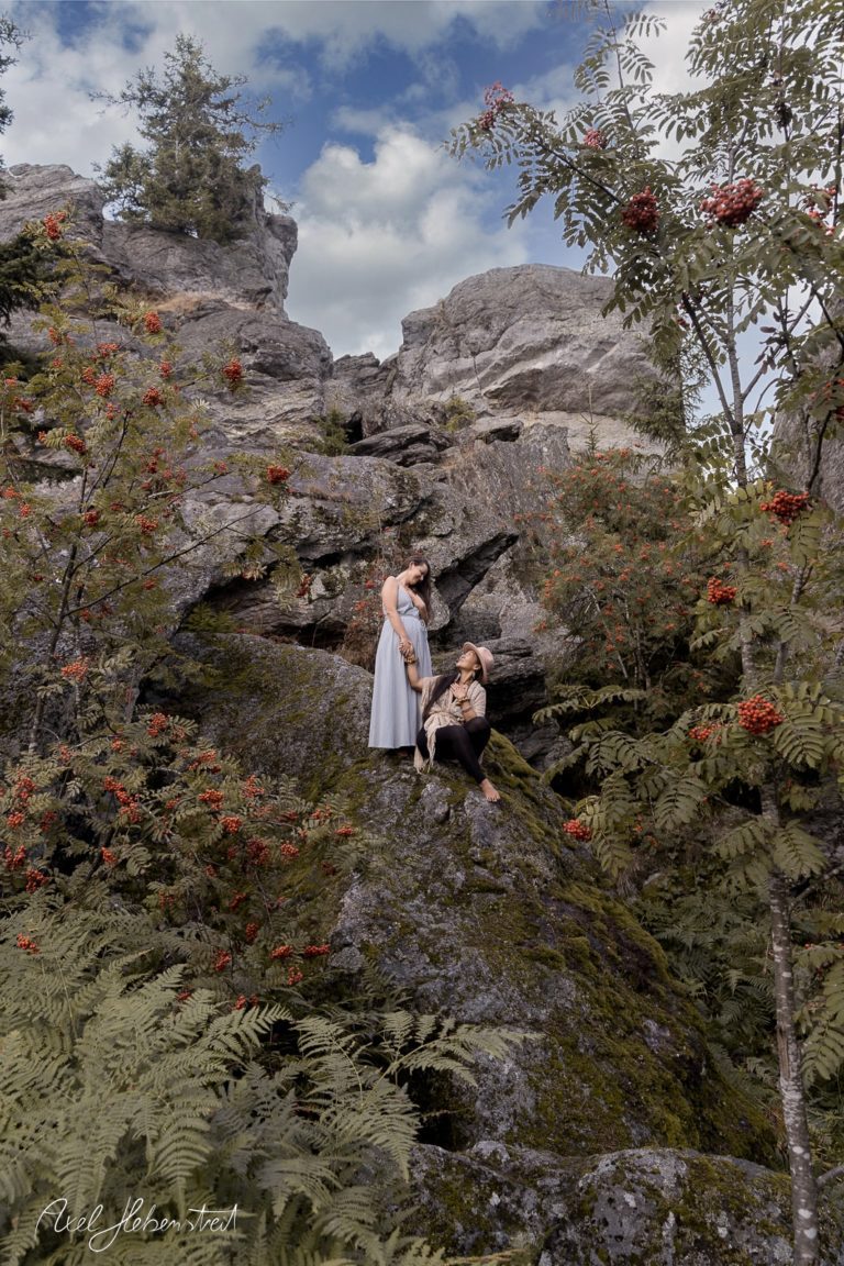 Zwei Frauen in eleganter Kleidung posieren auf einem Felsen umgeben von Bäumen und roten Beeren in der Natur – © Axel Hebenstreit.