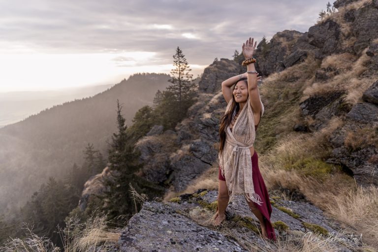 Eine Frau in traditioneller Kleidung steht auf einem Felsen, hebt die Arme zum Himmel und genießt die Natur. – © Axel Hebenstreit