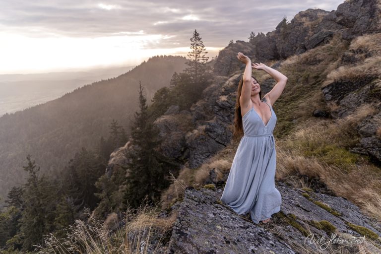 Eine Frau in einem langen, hellen Kleid tanzt auf einem felsigen Hang mit Blick auf eine bewaldete Berglandschaft – © Axel Hebenstreit