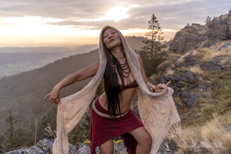 Eine Frau in traditioneller Kleidung posiert kraftvoll auf einem Berg mit einem dramatischen Sonnenuntergang im Hintergrund – © Axel Hebenstreit