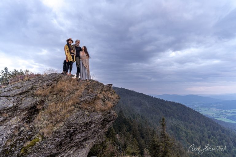 Drei Personen stehen auf einem Felsen mit Blick auf eine bewaldete Landschaft unter bewölkem Himmel – © Axel Hebenstreit