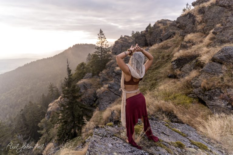 Eine Frau in einem weiten Rock mit gehobenen Armen steht auf einem Felsen und genießt die Aussicht in die Natur – © Axel Hebenstreit